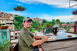 A student sits at a table outdoors on a balcony patio surrounded by building and green trees and plants. He holds a pen and has a notebook open on the table.