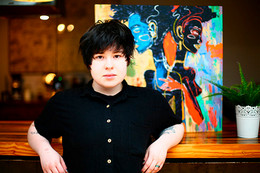 A student leans against a wood counter, propping their elbows up against the counter. A colourful painting is on the wall just behind them.