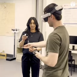 Two students stand in a lab. One student wears a virtual reality headset while the other student hands him the hand held remote.