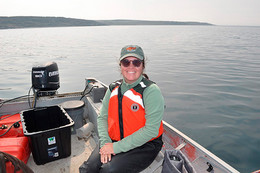 A student sits on a boat on the water with land seen behind her. She wears sunglasses, a baseball hat and a life jacket.